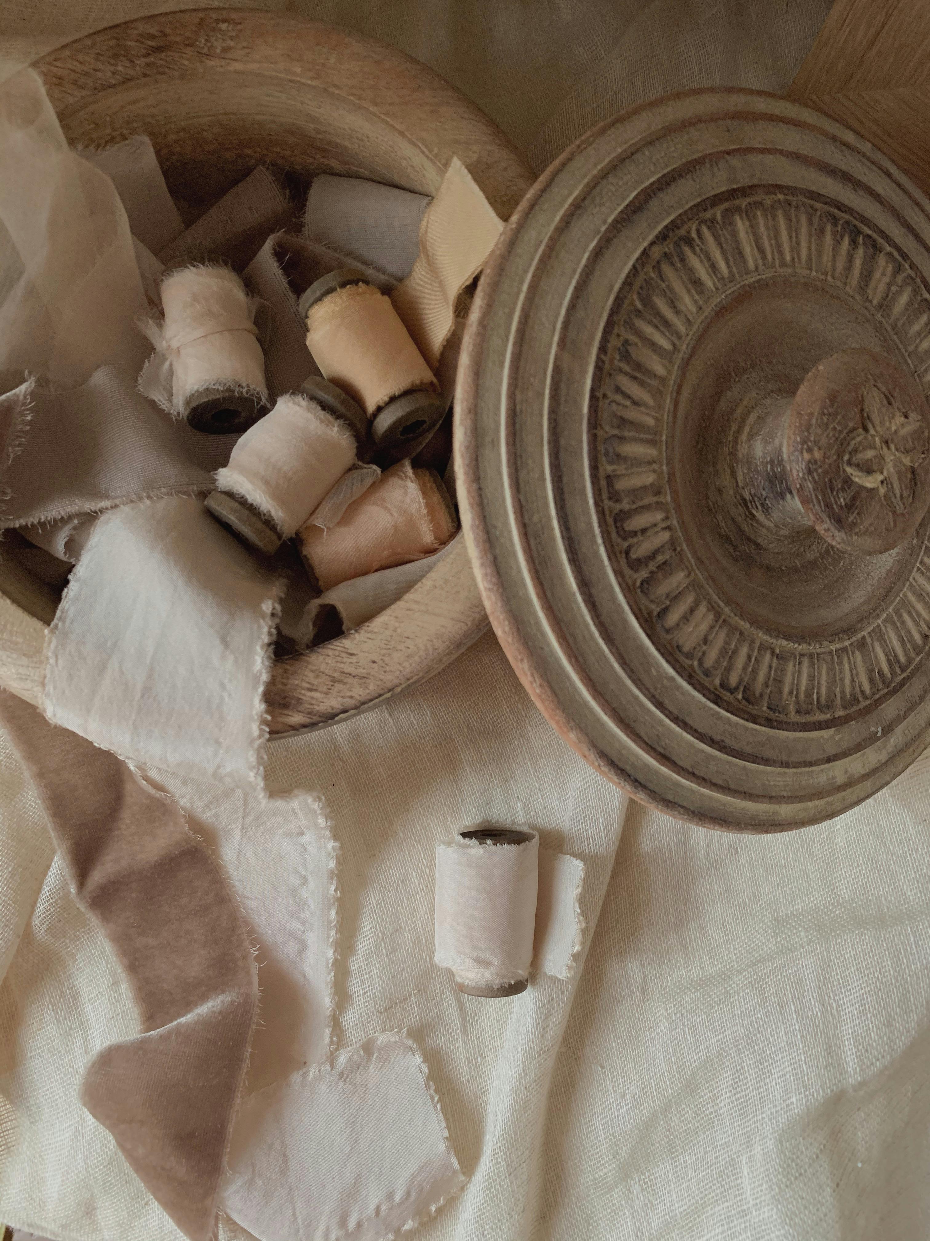 A vintage wooden bowl filled with neutral-toned spools of fabric ribbons, creating a rustic still life.