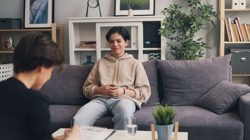 Free Teenager in therapy session sitting on a sofa, discussing with a professional therapist in a modern office setting. Stock Photo