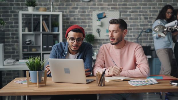 Two young professionals discussing work at a laptop in a modern office setting.