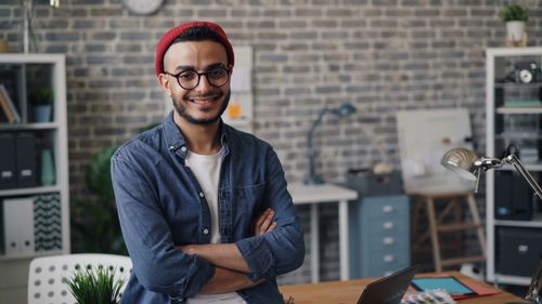 Free Smiling young man standing confidently in a modern office with a brick wall background. Stock Photo