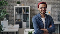 A man in glasses and a red hat is smiling in front of a desk