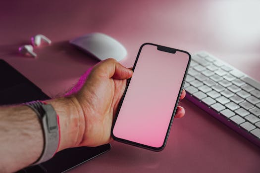 A hand holding a smartphone with a blank pink screen on a modern office desk.