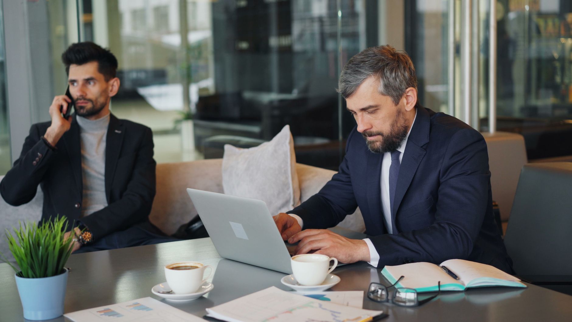 Two businessmen working in a modern office with laptops, coffee, and documents.
