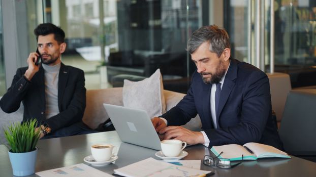 Two businessmen working in a modern office with laptops, coffee, and documents.