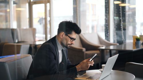 Free Man in formalwear using smartphone and laptop in a modern café setting. Stock Photo