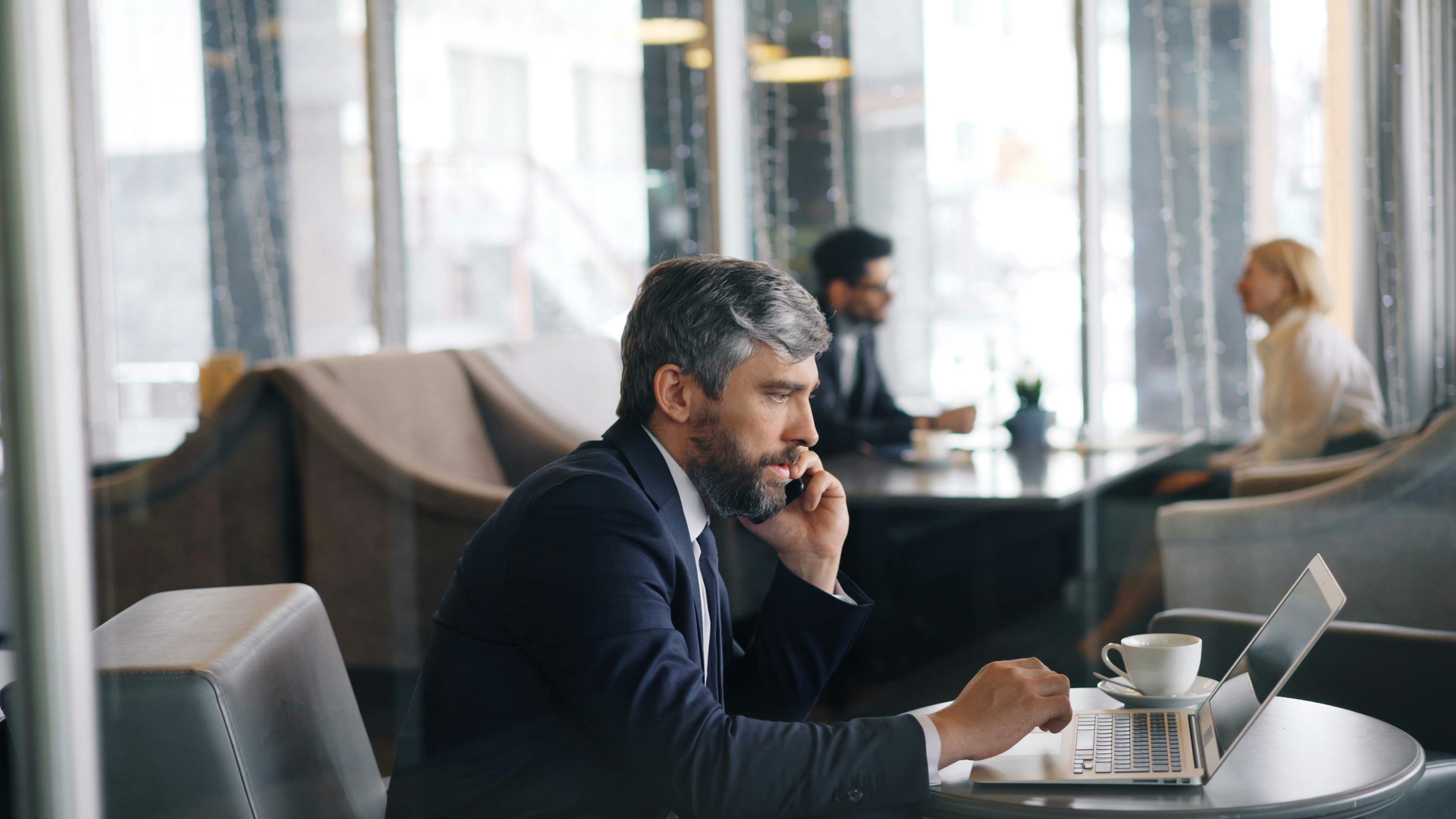 a man in a suit sitting at a table with a laptop and talking on the phone