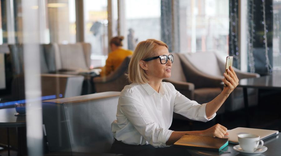 Woman making a video call on her smartphone at a cafe table with a coffee and laptop