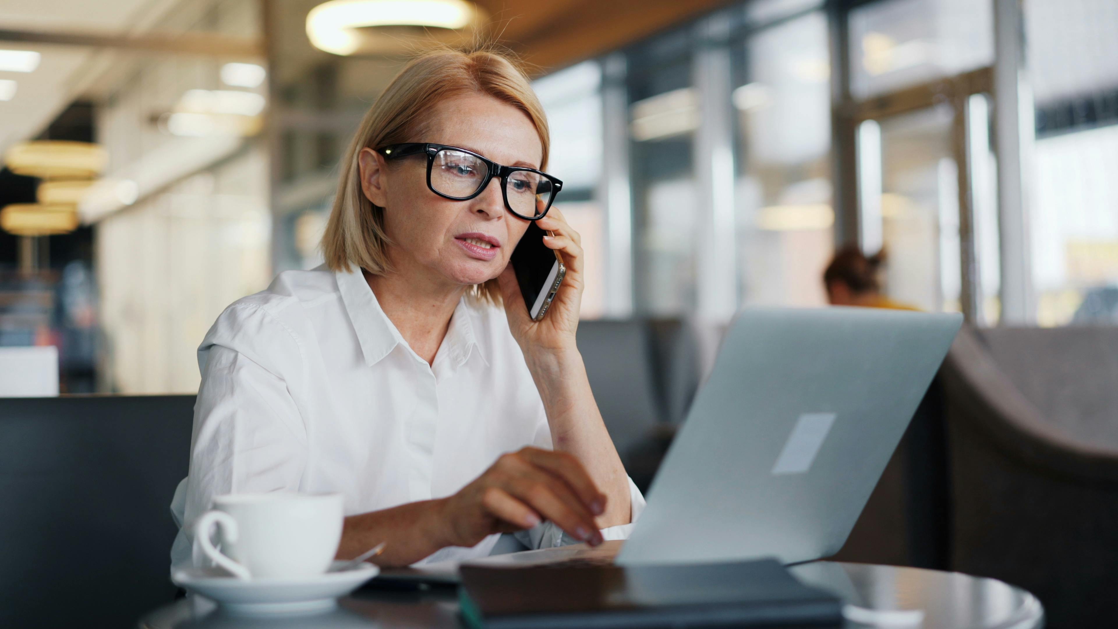 Mature woman in a café using a laptop and smartphone for business communication.