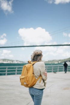 A woman walks by the Bosphorus Strait with a yellow backpack, viewing the iconic Istanbul bridge.