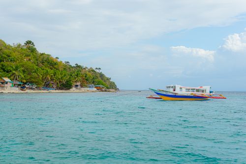 Free A vibrant boat floats near a lush green tropical coastline with palm trees and clear blue sea. Stock Photo