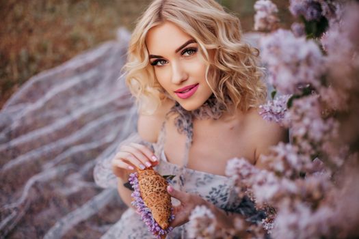 Blonde woman in a floral dress sits among lilac flowers, exuding elegance and charm.