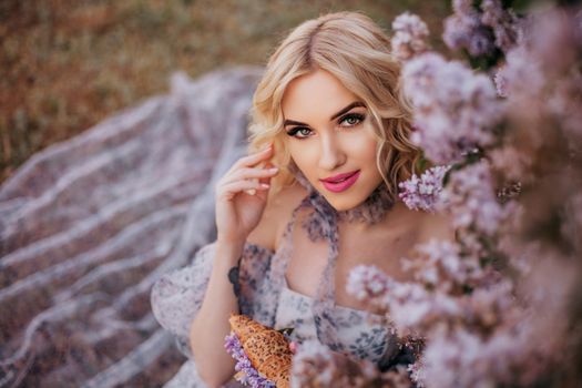Blonde woman in a tulle dress posing elegantly among lilac flowers in a garden.