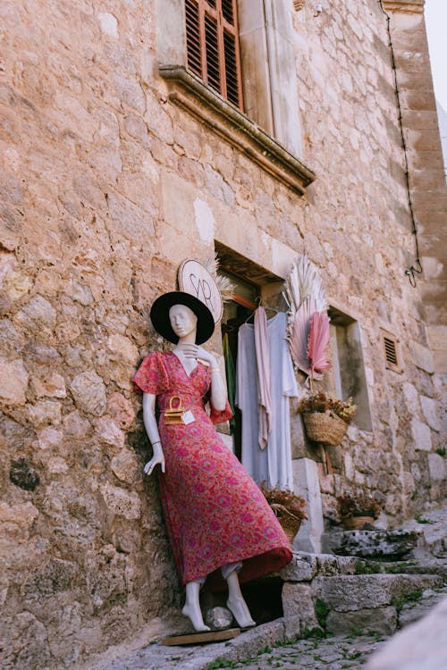 A mannequin dressed in a pink dress stands outside a stone building