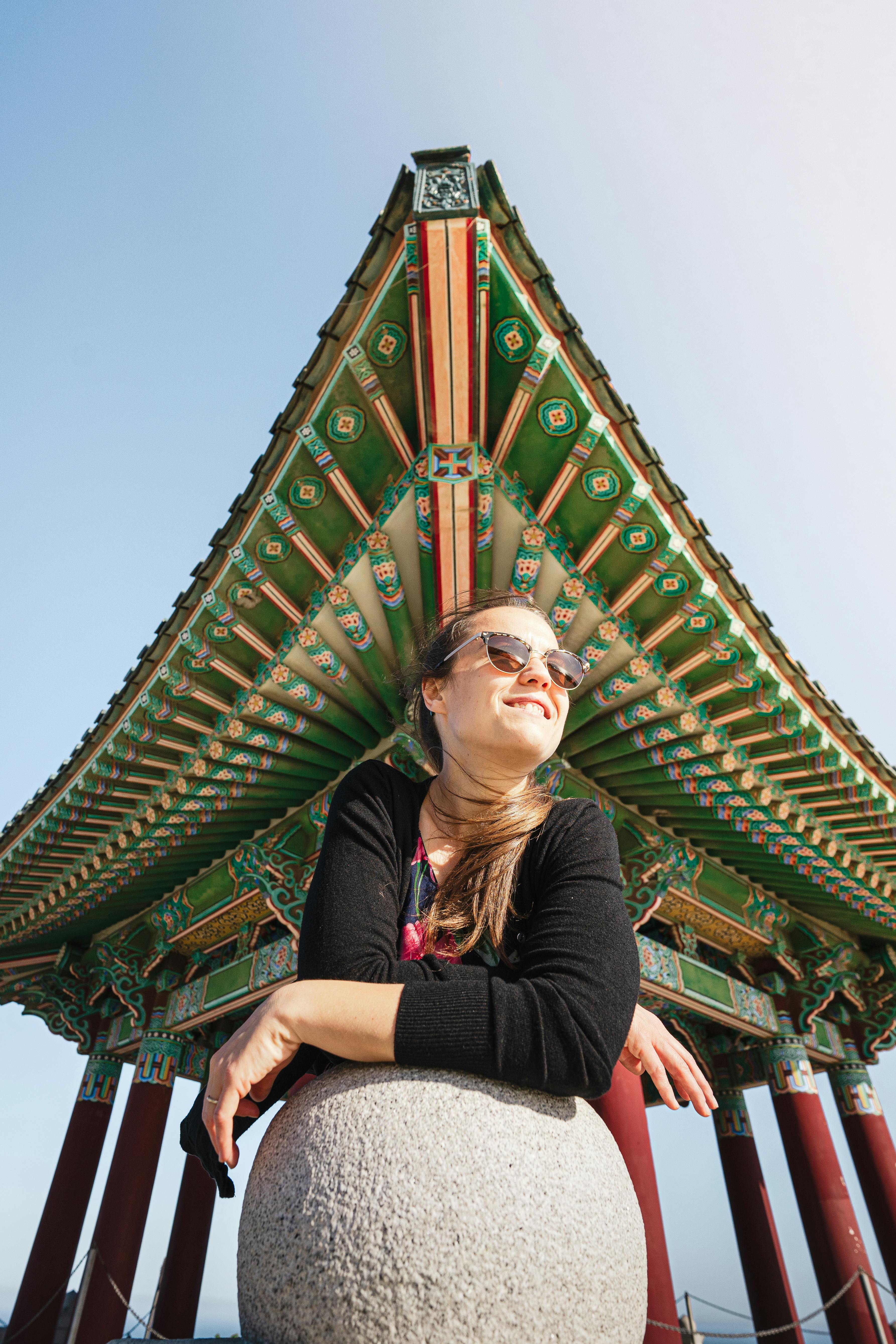 Smiling woman leaning on stone ball beneath colorful Korean pavilion.
