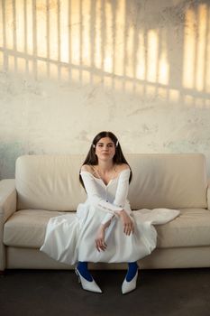 Stylish woman in a white dress and blue heels sitting on a sofa under soft lighting.