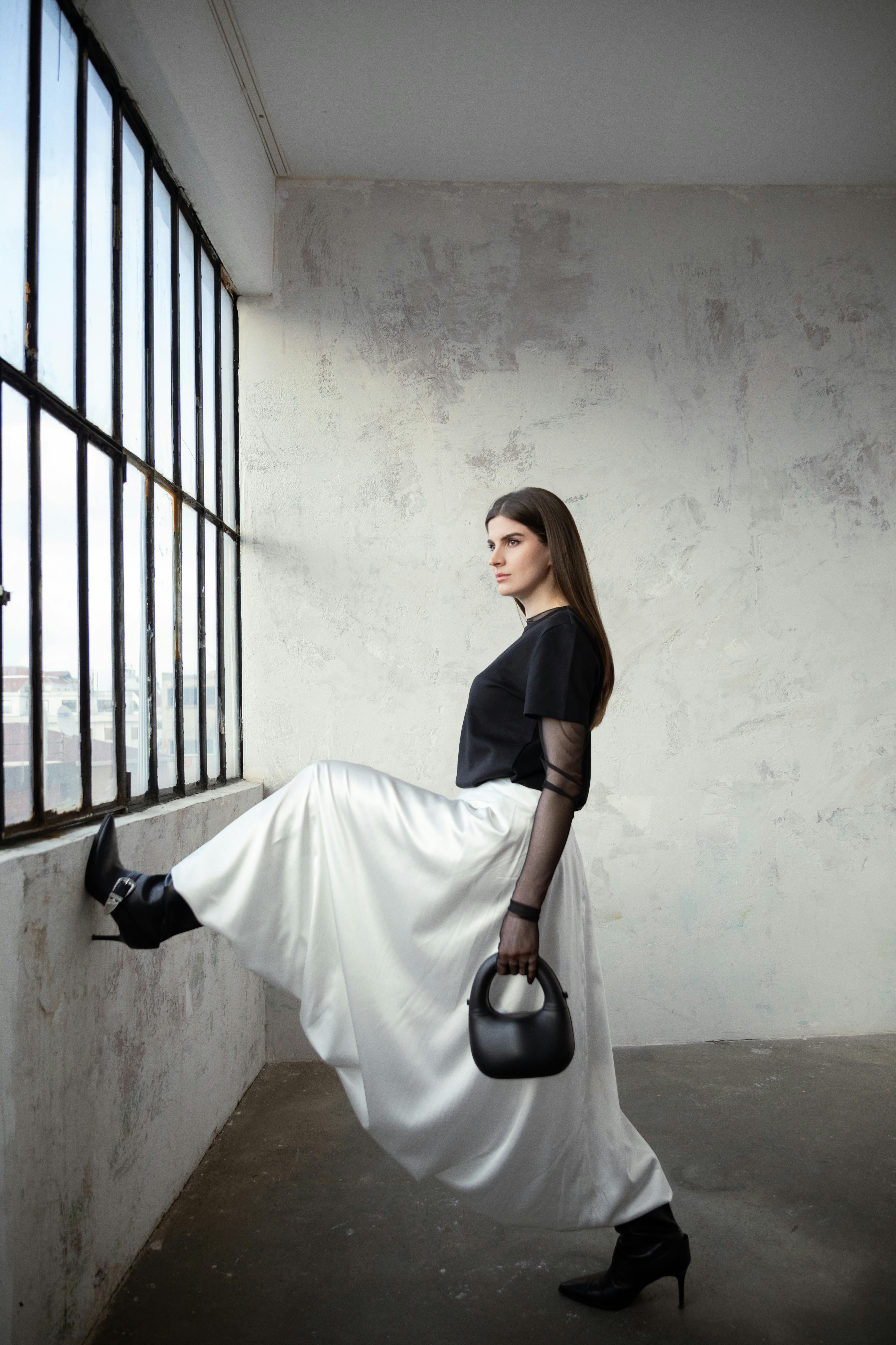 Stylish woman in black top and white skirt posing by large window in industrial setting.