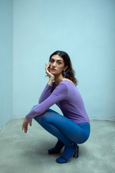Elegant fashion model posing in vibrant purple bodysuit and blue tights indoors.