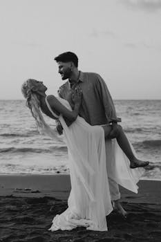 Elegant couple in romantic embrace by the sea during twilight, captured in black and white.