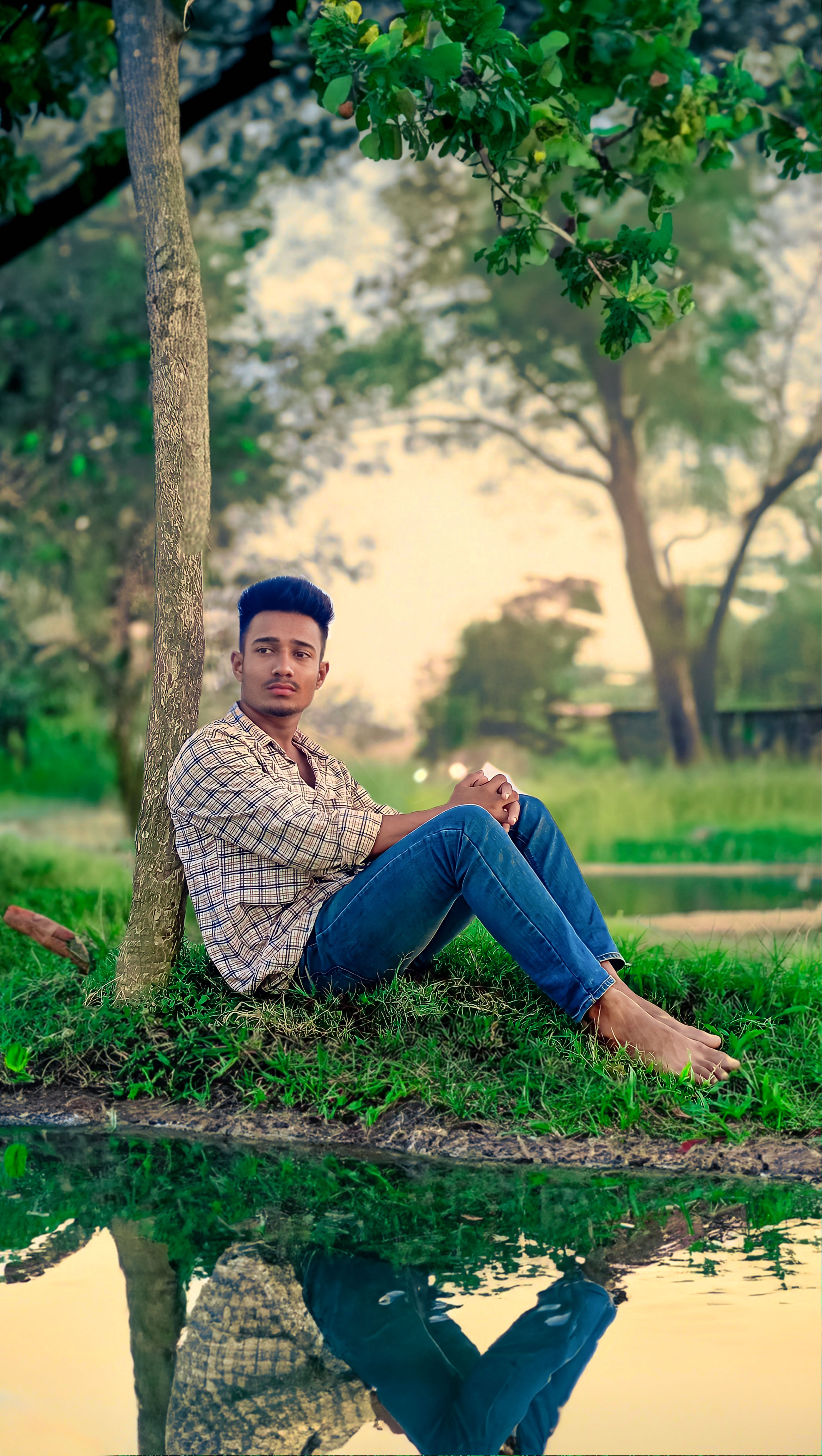 Young man sits by a tree beside a reflective pond, enjoying a peaceful outdoors setting.