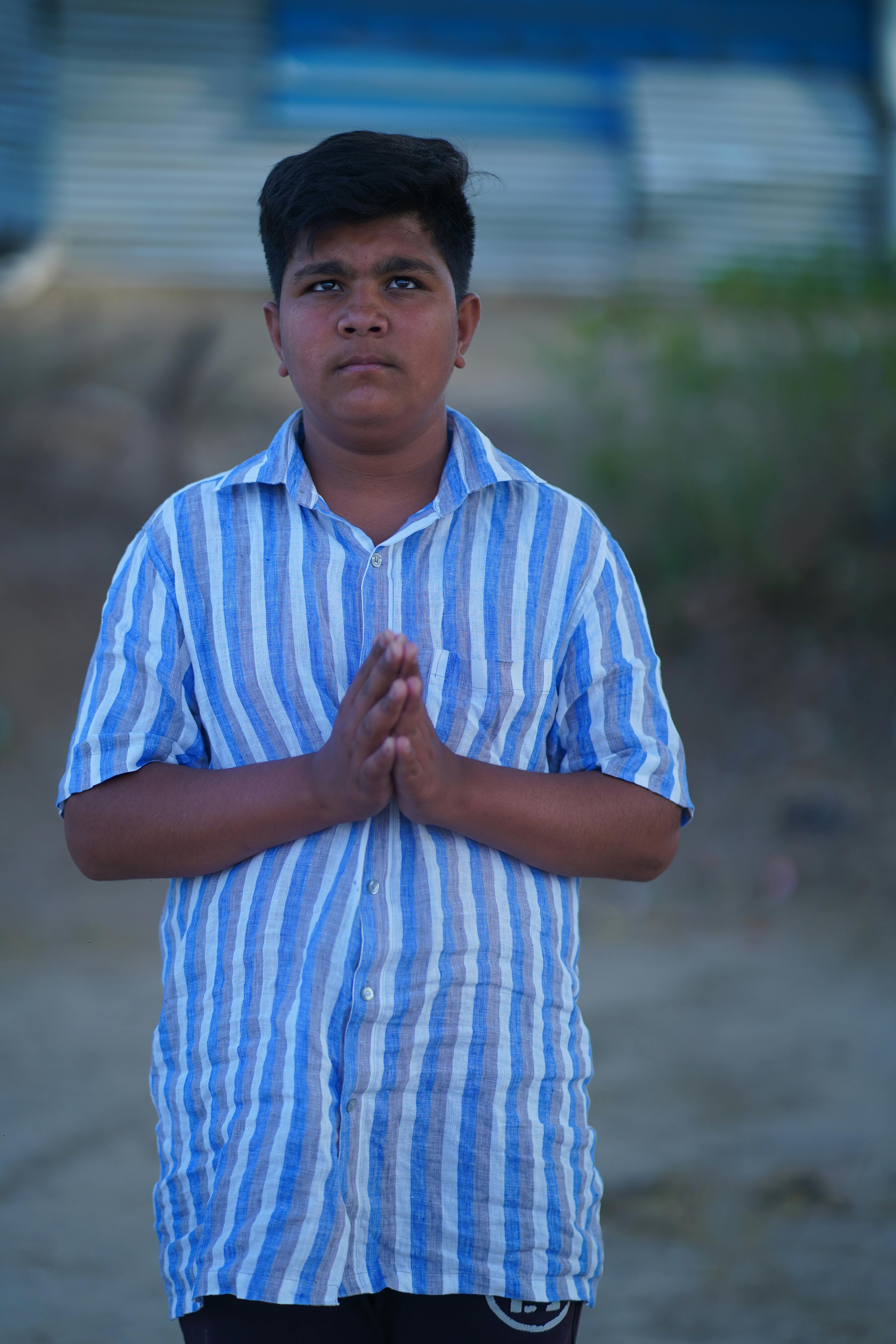Teenage boy in striped shirt stands outdoors with hands in prayer, exuding a sense of spirituality.