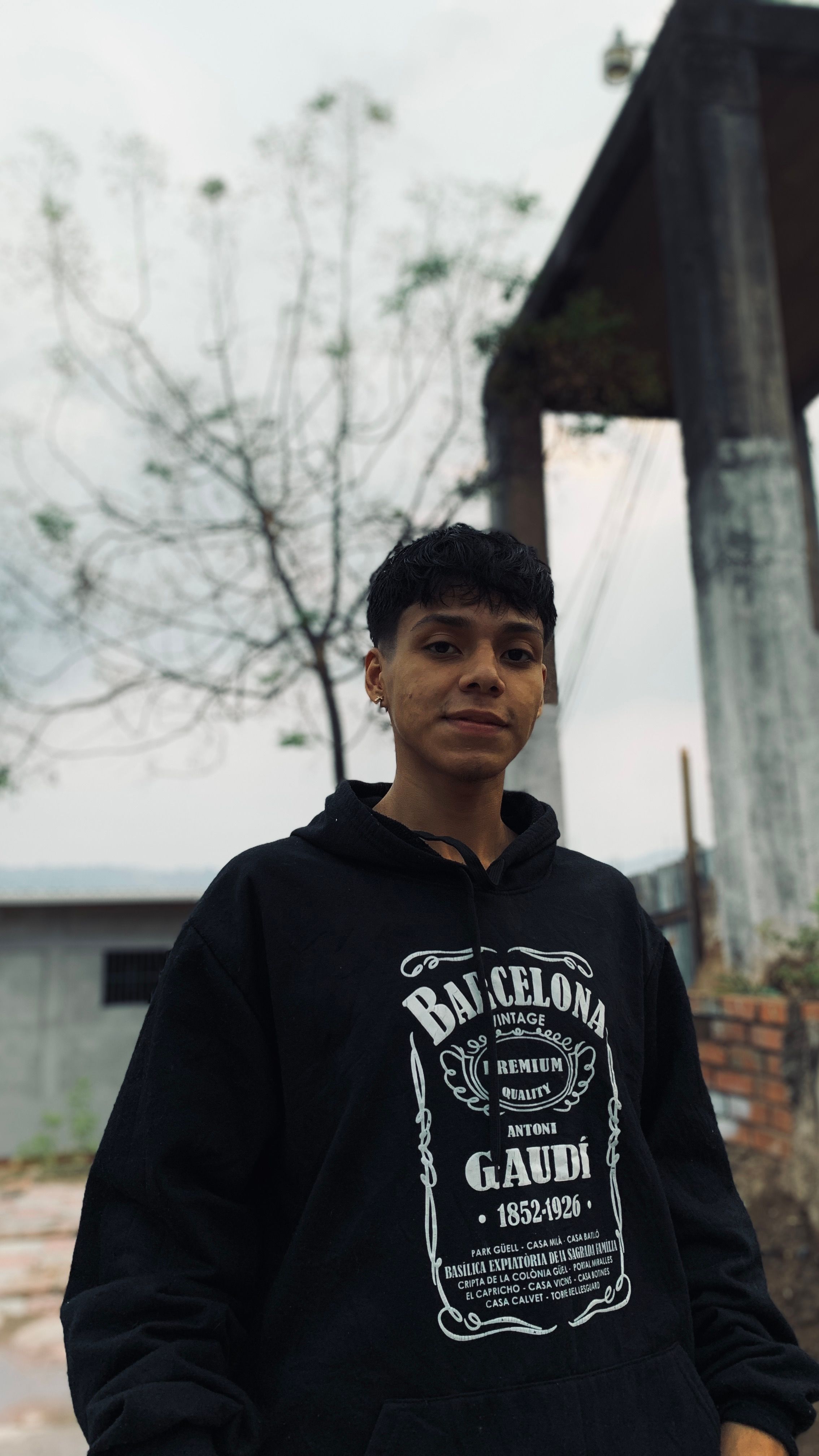 Young adult in black hoodie with Barcelona text standing outside with trees and concrete structure.