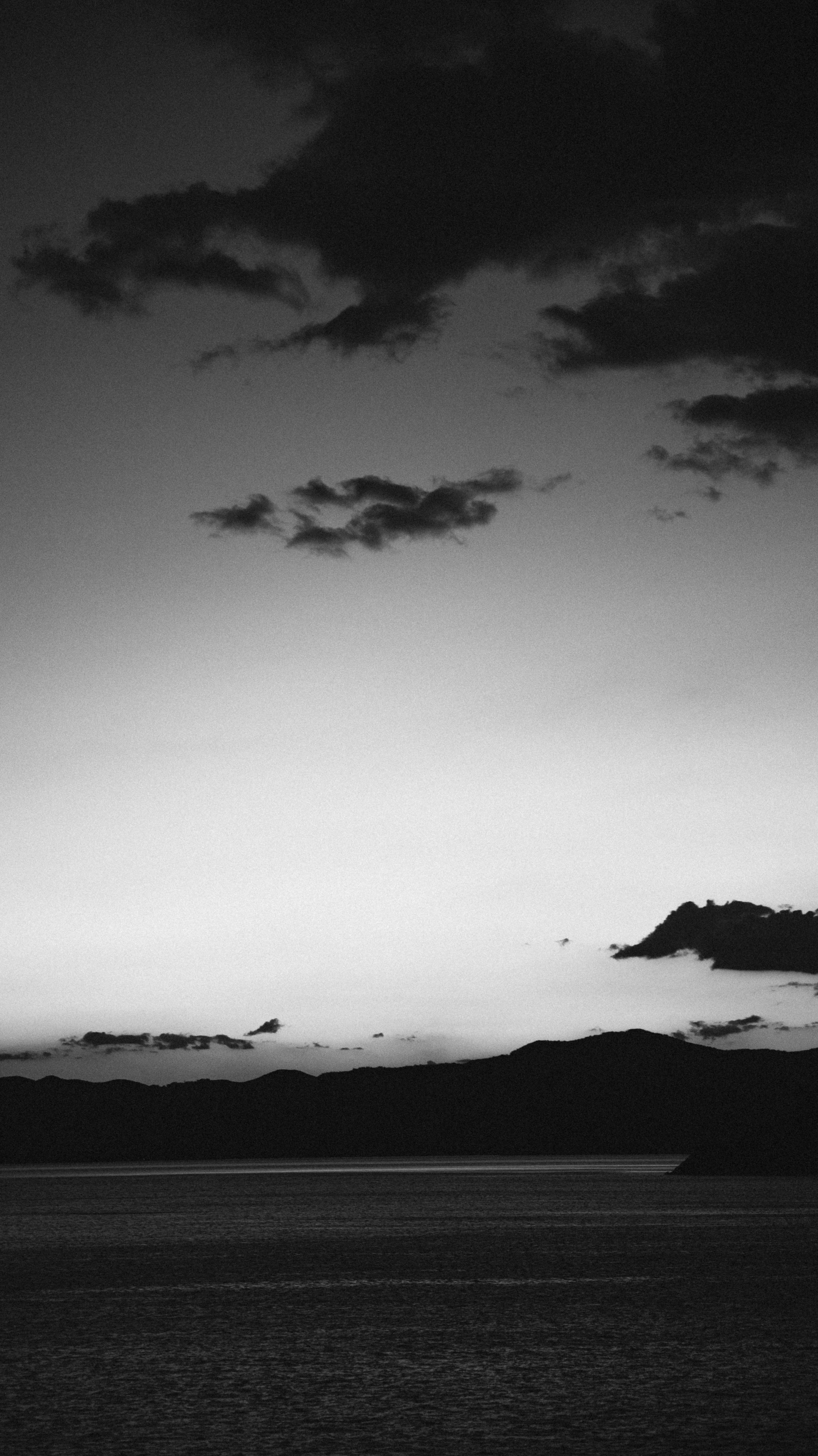 Monochrome coastal scene capturing silhouetted hills against the cloudy evening sky.