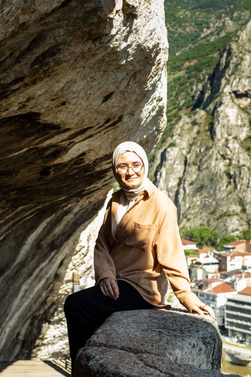 Free A smiling woman in a hijab sitting under a rock ledge enjoying the sunlight in a scenic mountainous area. Stock Photo