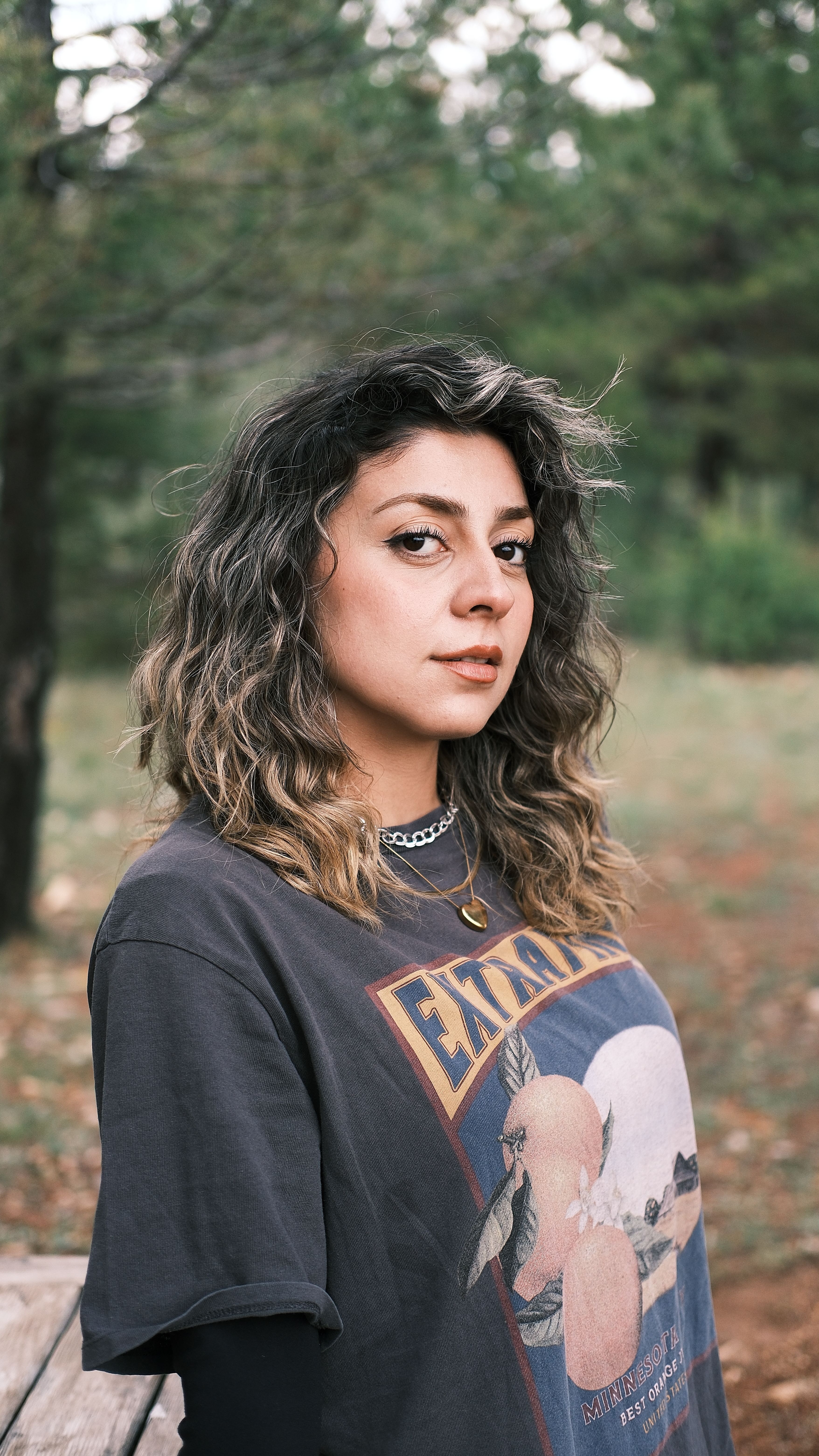 A stylish young woman with curly hair poses outdoors amidst trees, showcasing casual fashion.