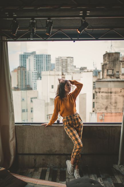 Trendy woman in orange sweater posing against cityscape backdrop indoors.