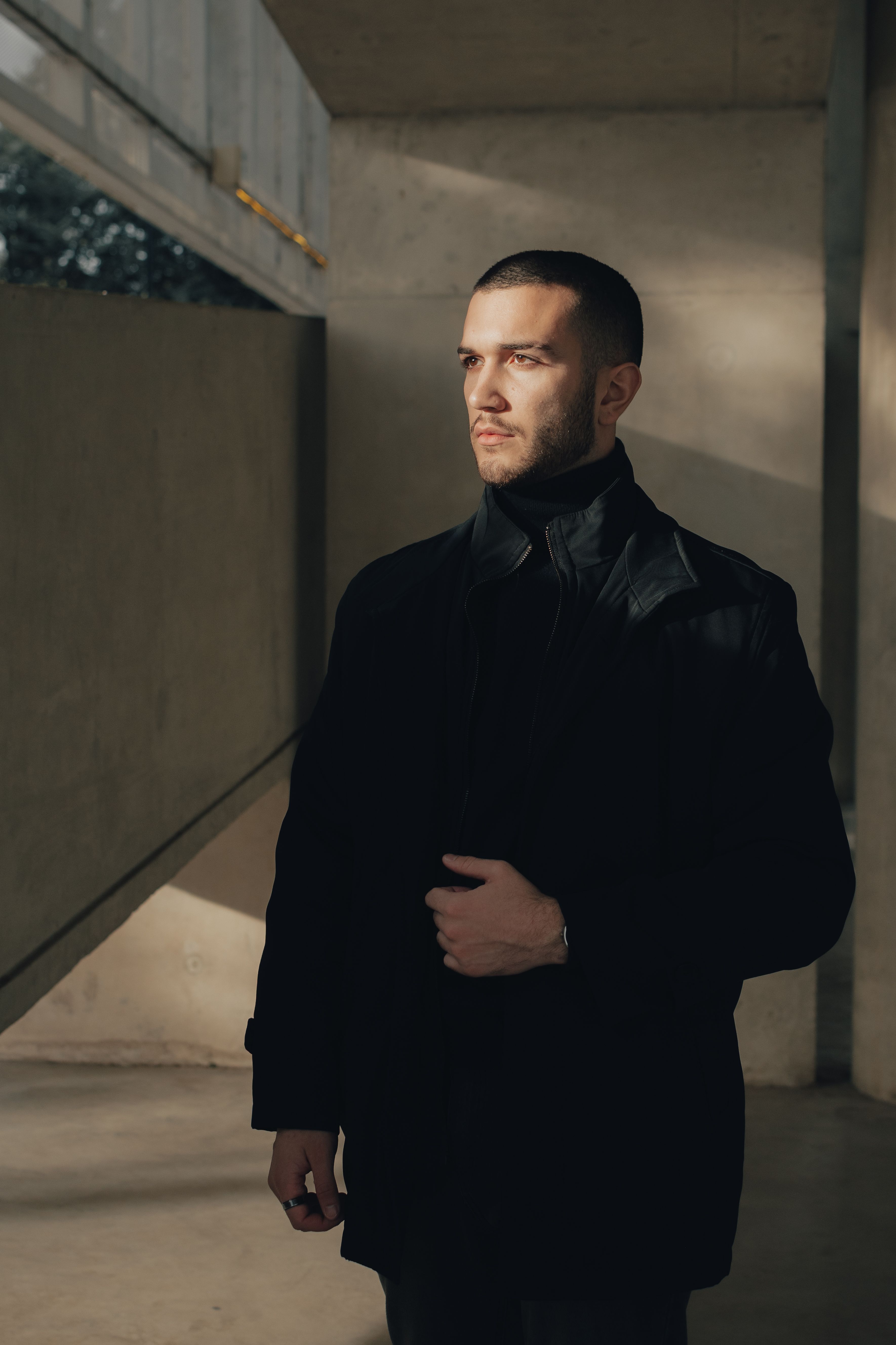 A well-dressed man in black clothing standing in a shadowy indoor setting.