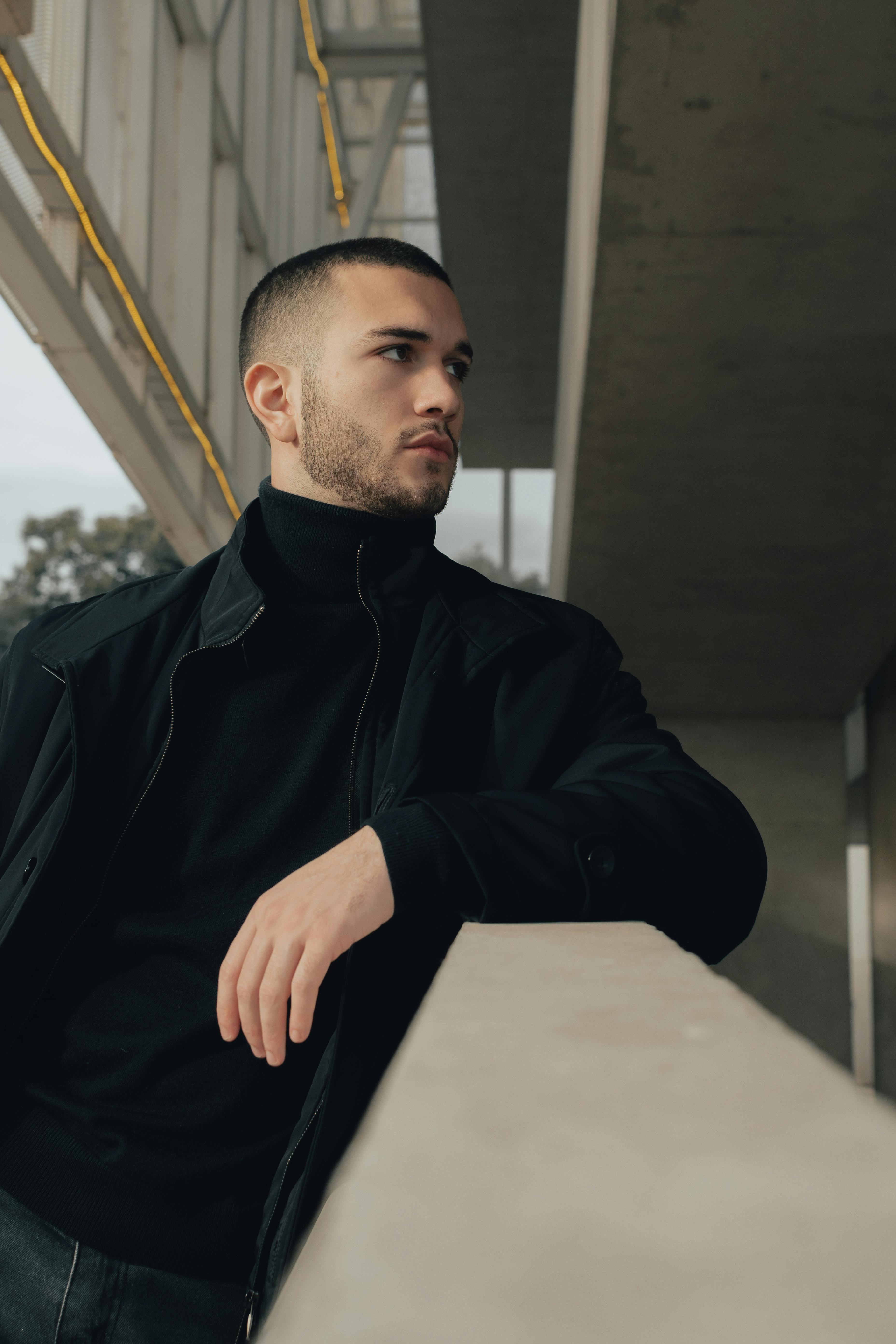 A fashionable man in a black outfit poses thoughtfully in an urban city environment.