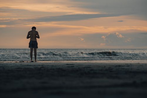 Free A lone figure stands on the beach, silhouetted against a vivid sunset sky. Stock Photo
