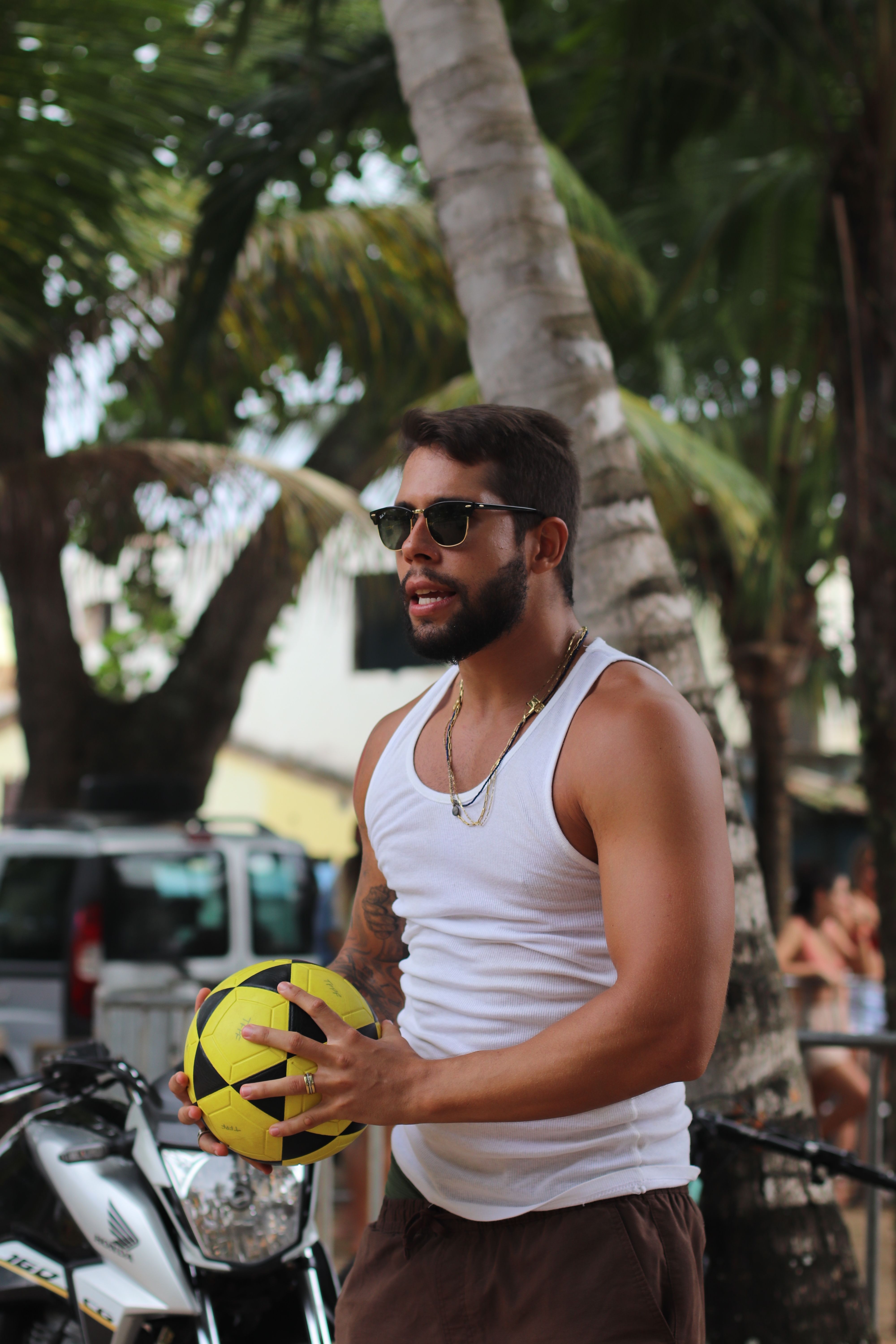 A man in sunglasses holds a yellow soccer ball outdoors near palm trees.