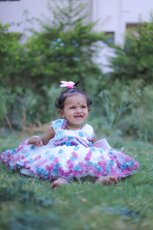 Free Cute baby girl in a colorful dress sitting in a park, smiling joyfully in the greenery. Stock Photo