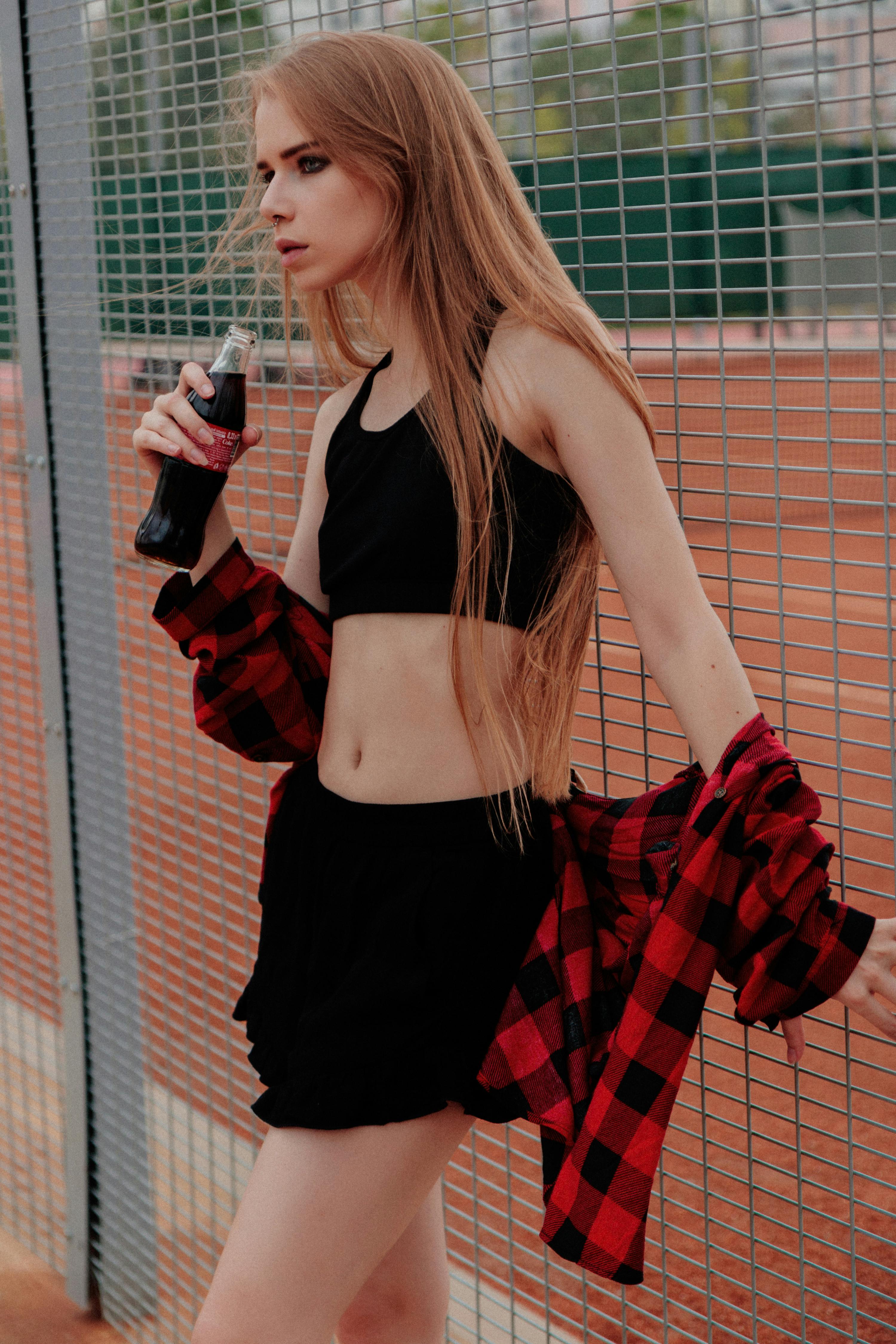 Young woman in sporty attire holding a drink, leaning against a fence in an outdoor setting.