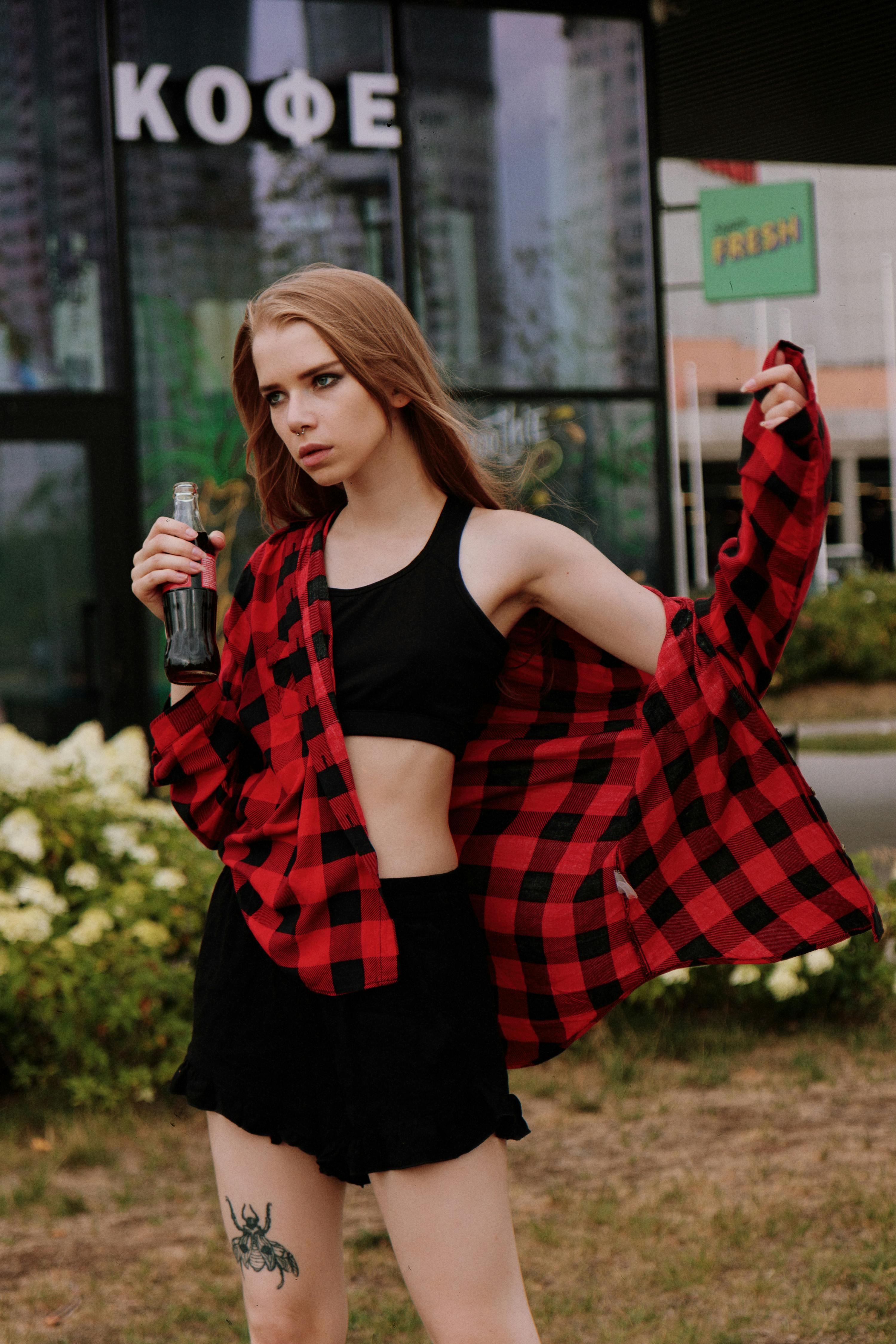 Stylish young woman posing outdoors with a soda bottle, showcasing tattoos and modern fashion.