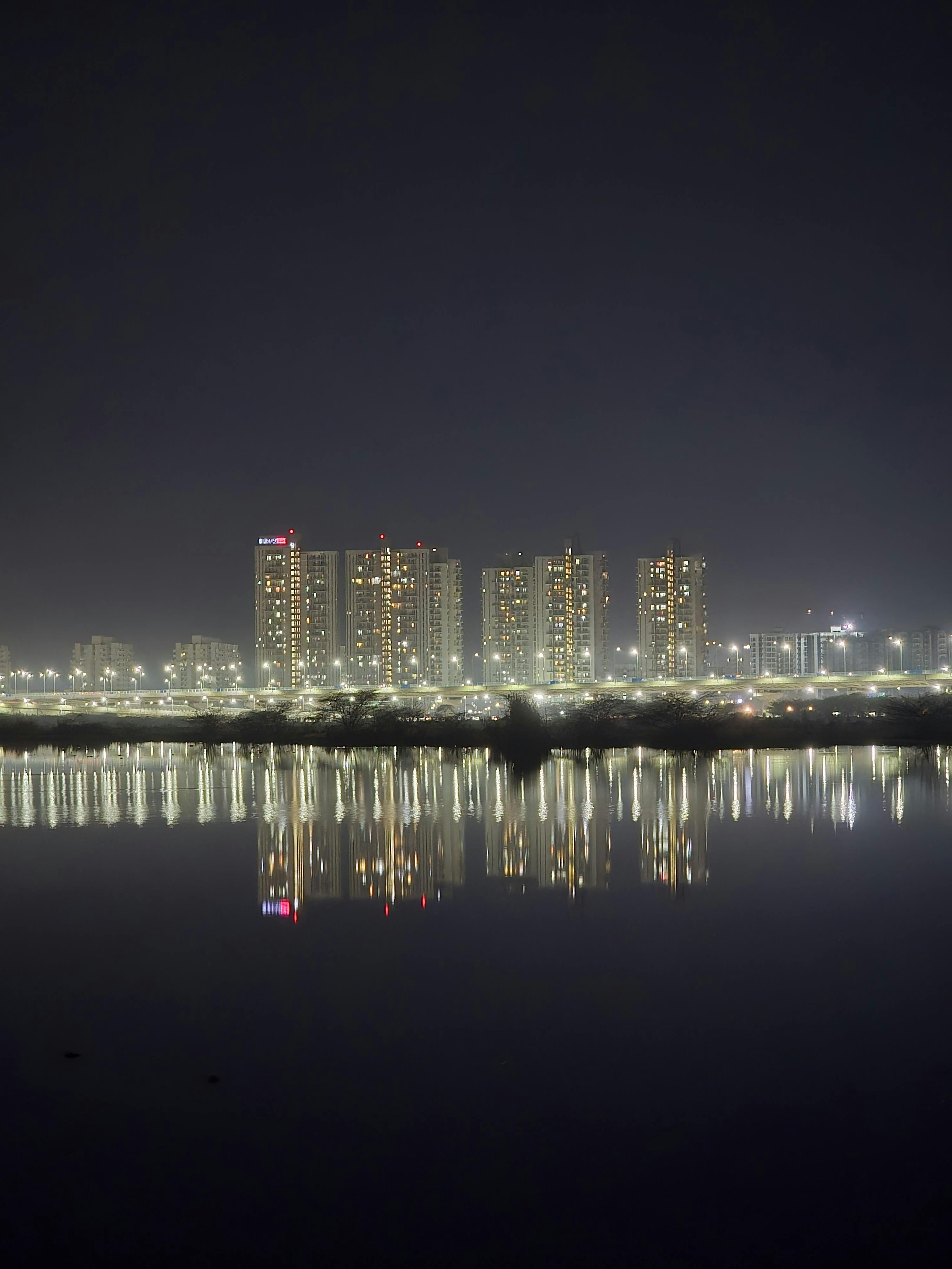 Bright city skyline of Gurugram with skyscrapers reflecting on a calm river at night.
