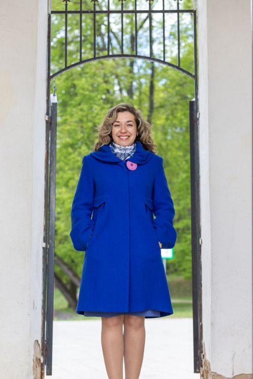 Free Elegant woman in a vibrant blue coat standing and smiling at a gate in a spring setting. Stock Photo