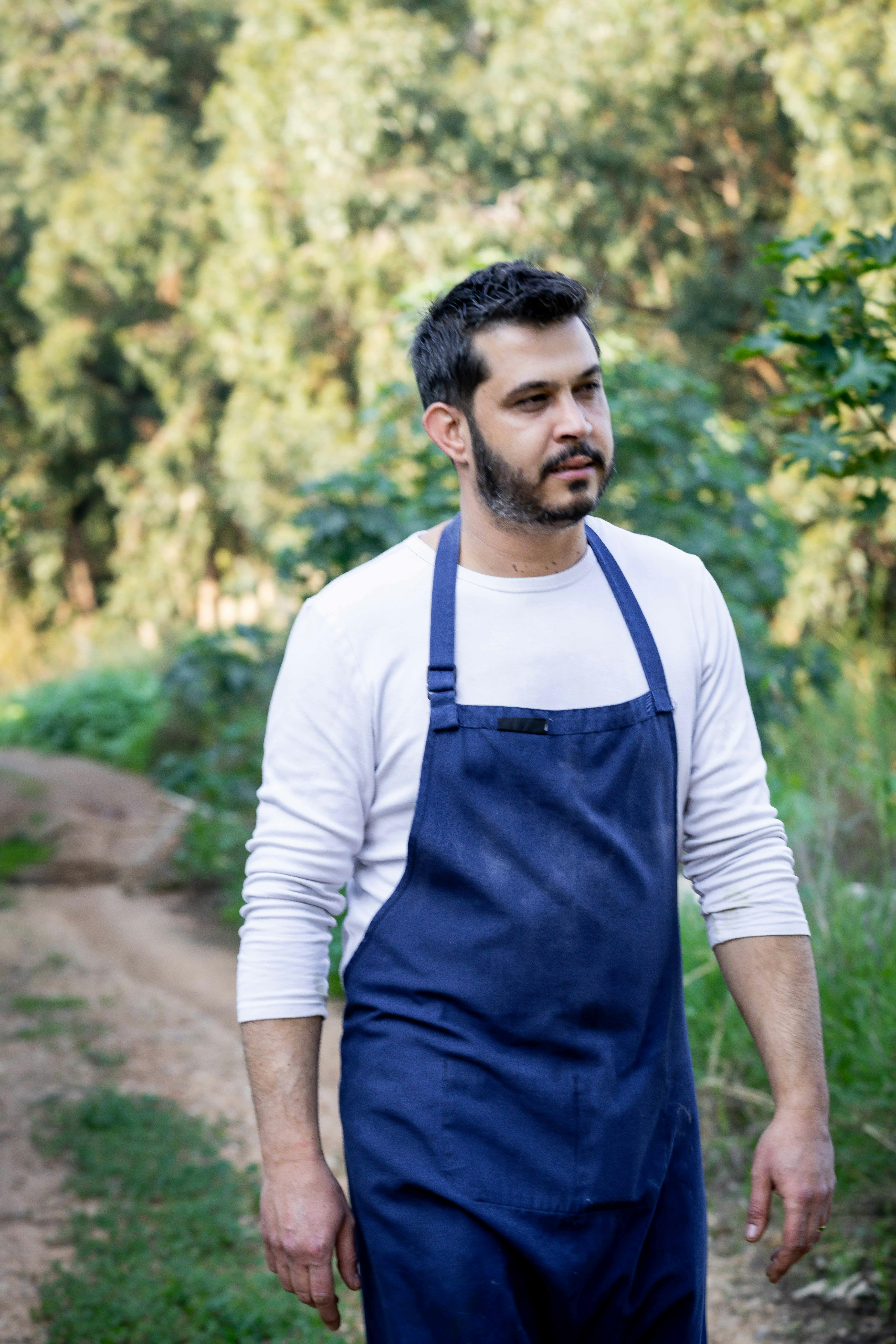 Man in blue apron walks through lush rural field, surrounded by trees and greenery.