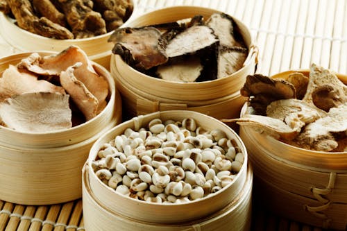 From above many wooden containers filled with assorted dried mushrooms with condiments and raw cashew nuts placed on bamboo mat