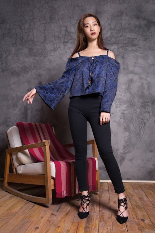 Woman in Blue Off-shoulder Top Standing Near Brown Wooden Rocking Armchair
