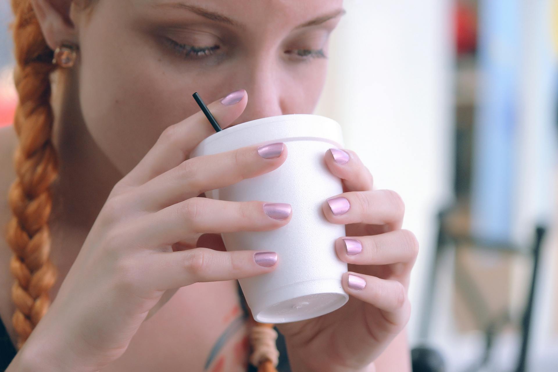 A young woman with braids enjoys a warm drink from a styrofoam cup indoors.