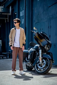 Stylish young man in sunglasses stands next to a motorcycle on a city street.