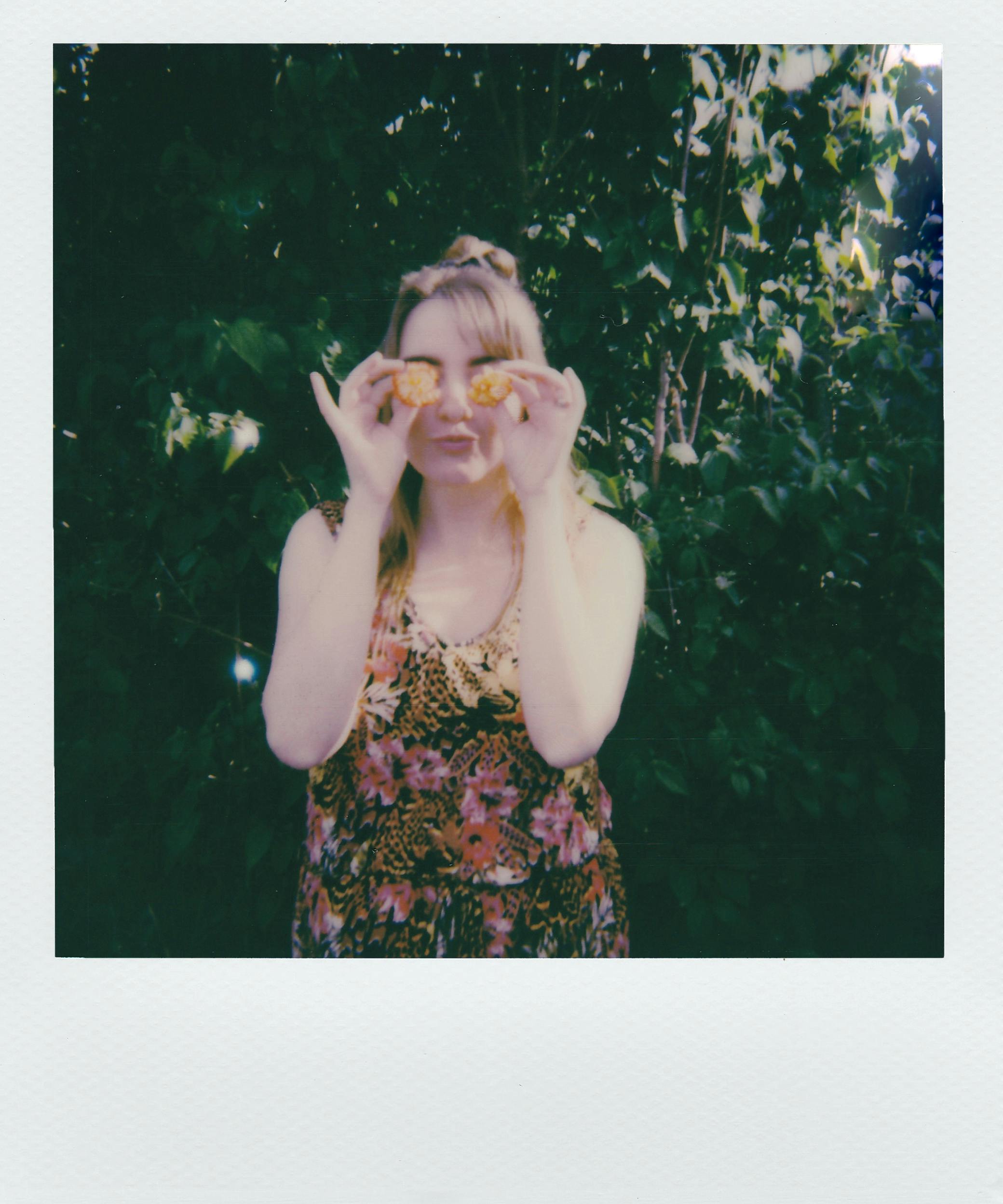 A woman playfully holds flowers in front of her eyes, surrounded by lush greenery.