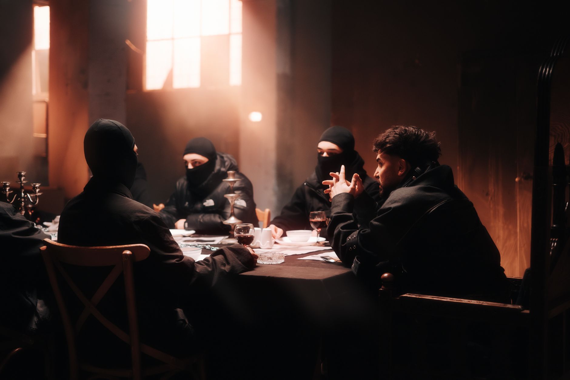 Men in dark attire with balaclavas gather around a table in a shadowy room with sunbeams filtering in.