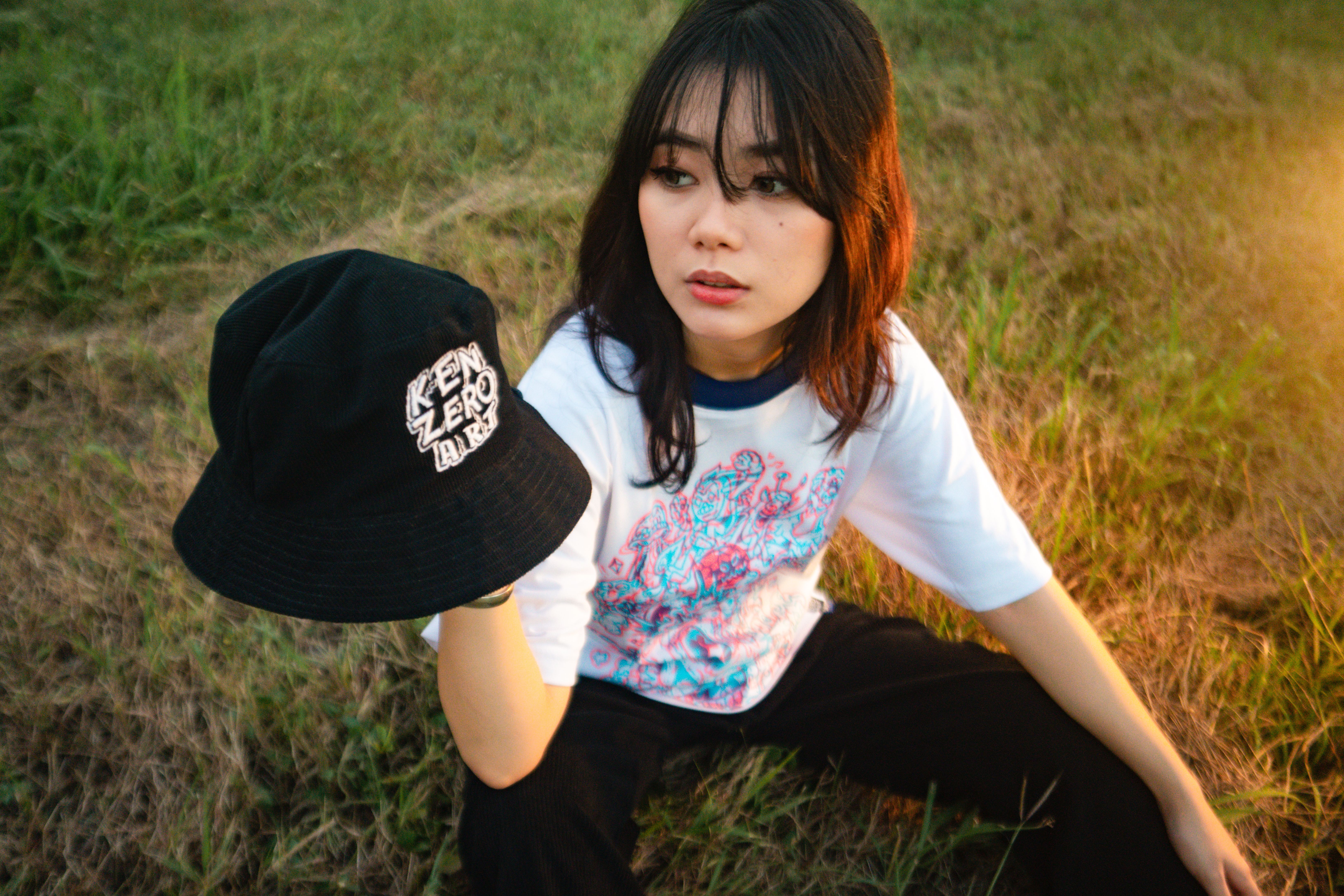 Woman sitting on grass outdoors holding a stylish black hat during sunset.