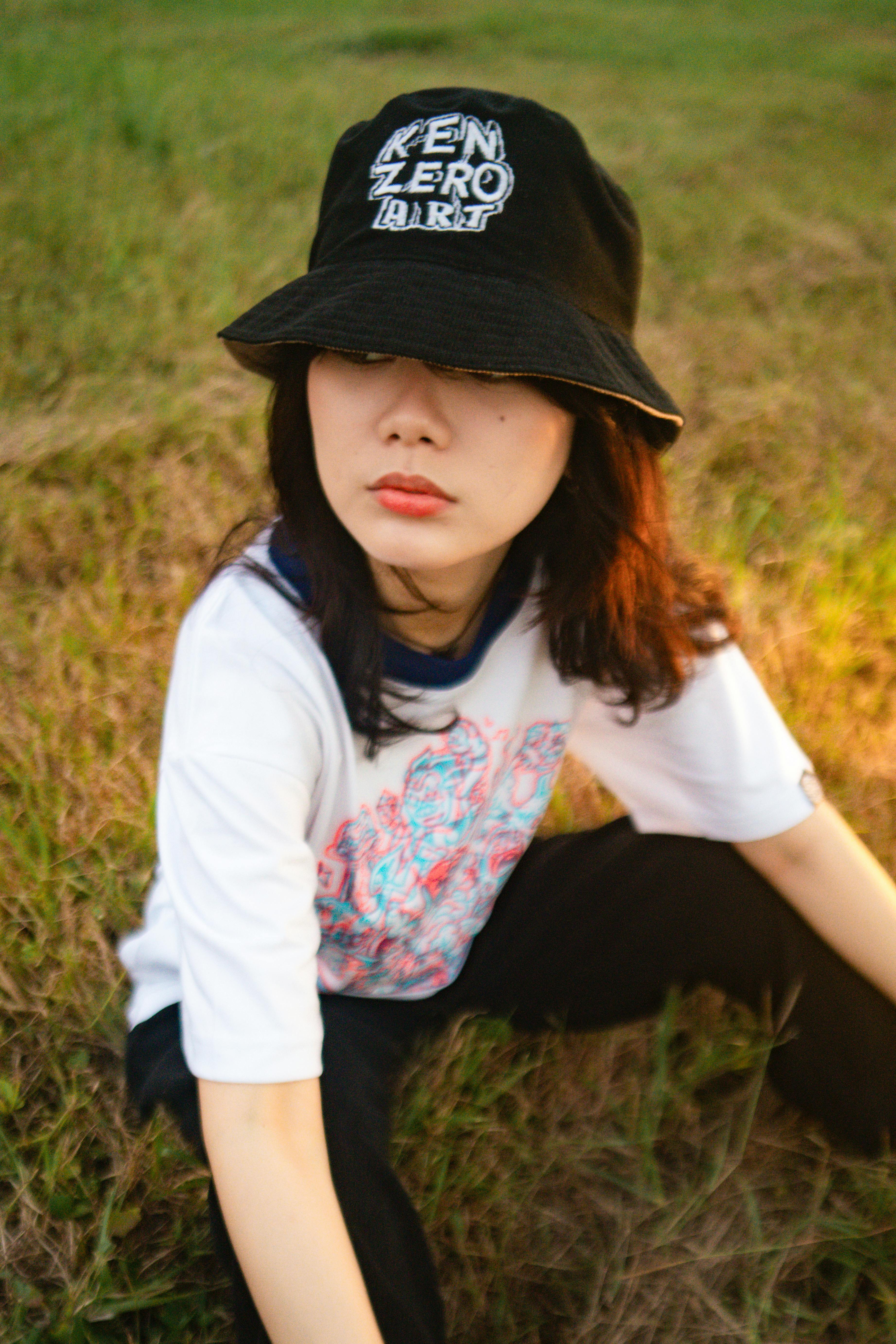 Portrait of a fashionably dressed woman wearing a hat, sitting on grass in a rural setting, creating a relaxed vibe.