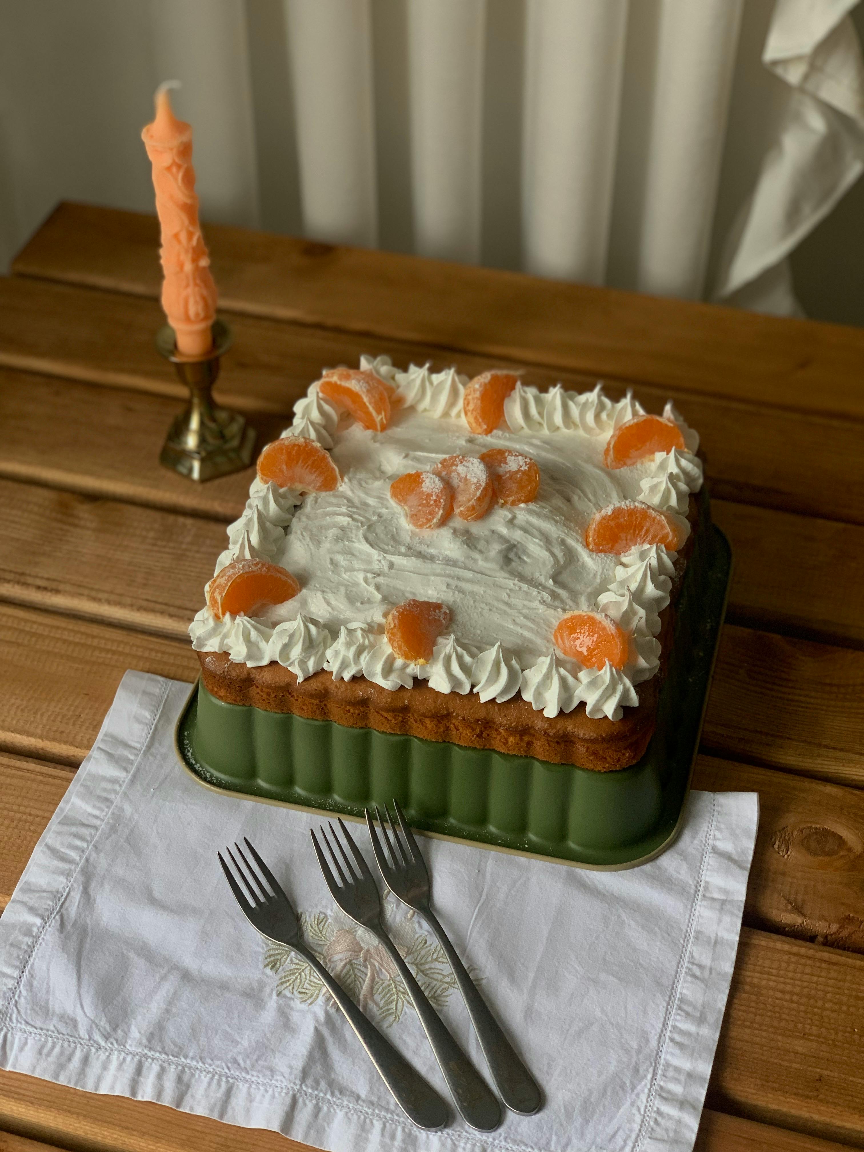 Square cake with whipped cream and tangerines on wooden table, next to a candle.
