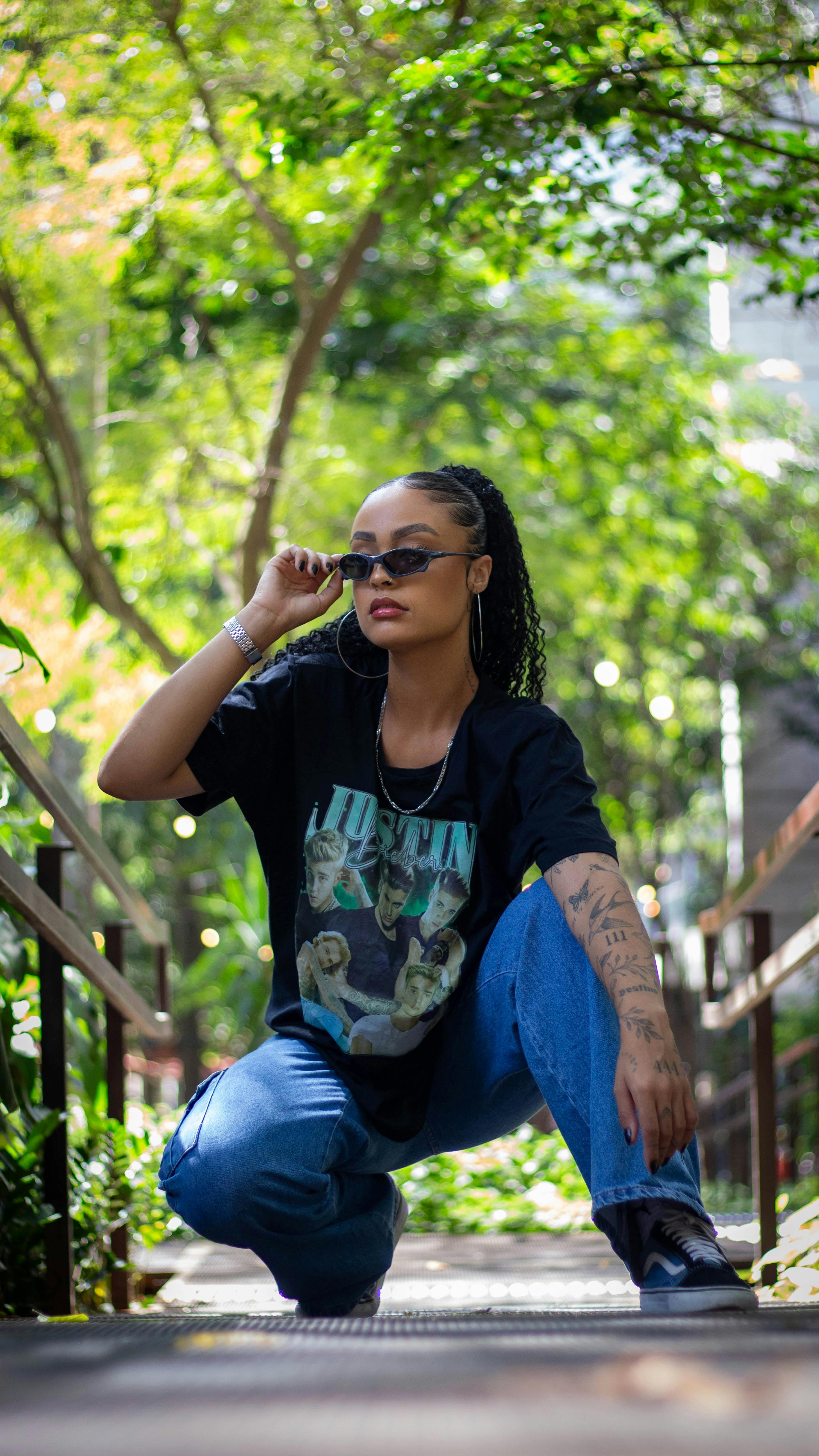 Stylish woman crouching with sunglasses in a lush outdoor setting during summer.