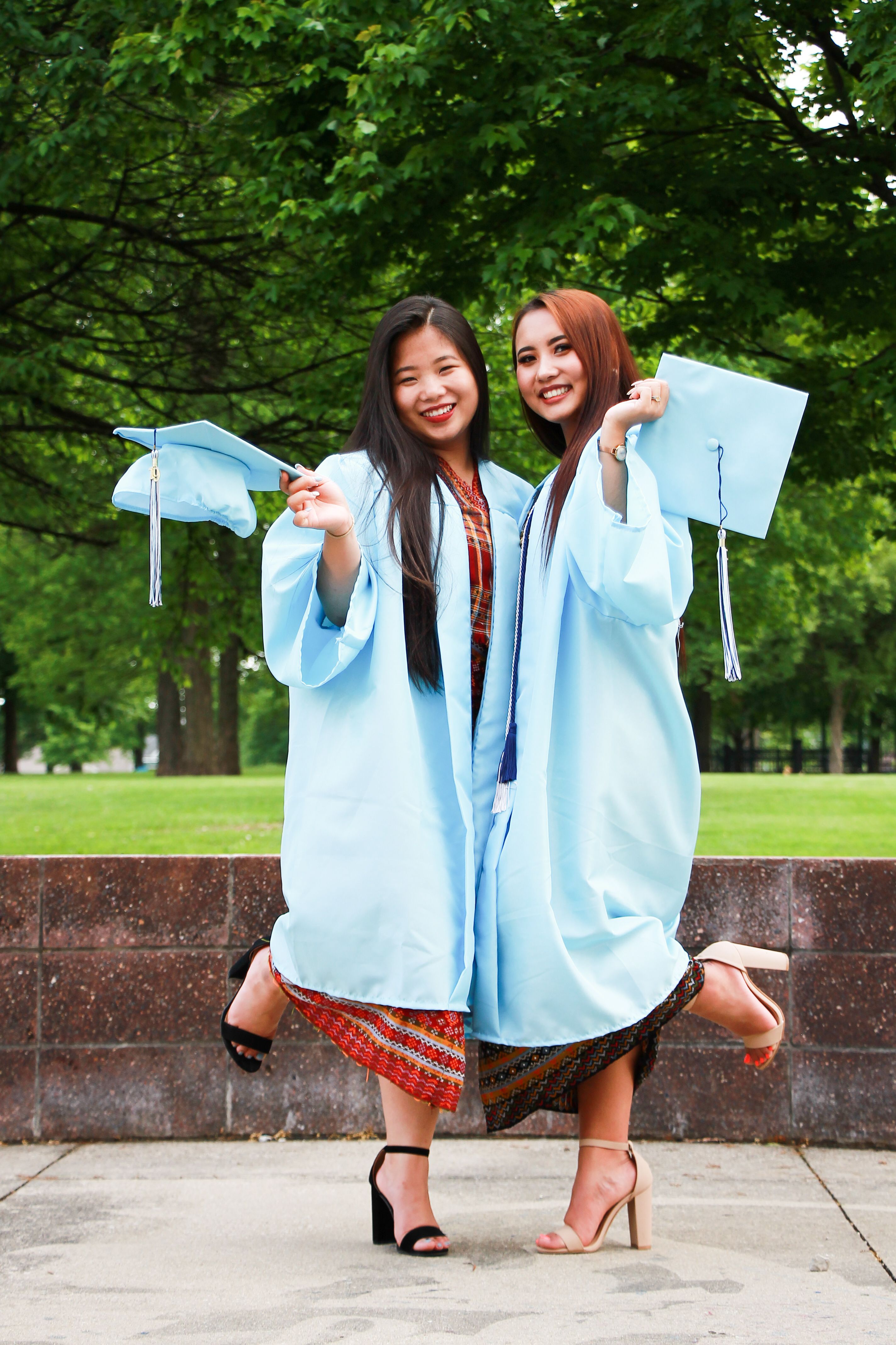 Two smiling young women wearing light blue graduation gowns and holding caps in a green park